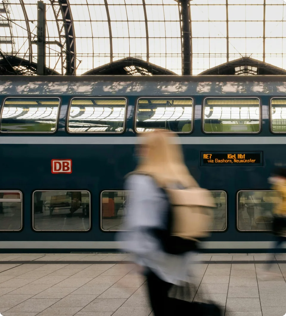 Un tren de dos pisos de Deutsche Bahn en el andén de una estación bajo una cubierta de cristal, con un viajero que pasa desenfocado por el movimiento